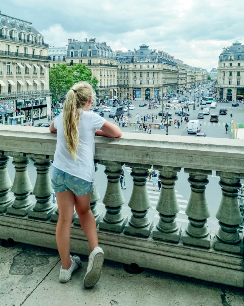 Foto van meisje dat van het balkon van Opéra Garnier uitkijkt over de Place de l'Opéra in Parijs.