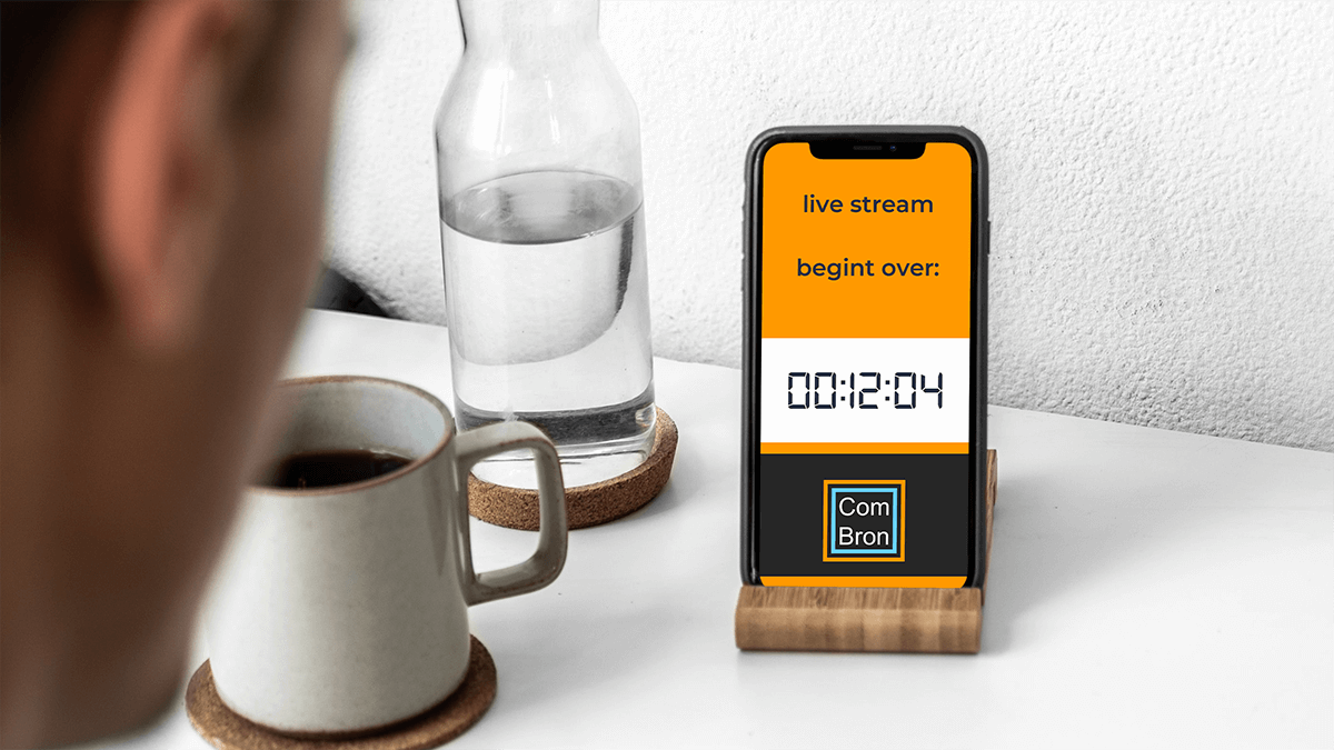 Mockup van een man aan een bureau. Op zijn telefoon staat te lezen: "live stream begint over 12:04 ComBron".